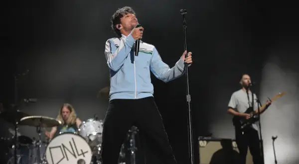 Louis Tomlinson na scenie, śpiewający do mikrofonu. W tle perkusista i gitarzysta.