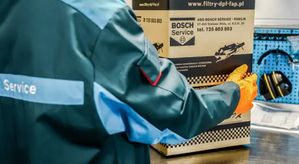 Pracownik w pomarańczowych rękawiczkach trzyma karton z filtrem DPF. Dowiedz się, ile trwa wypalanie dpf podczas jazdy, aby uniknąć problemów.