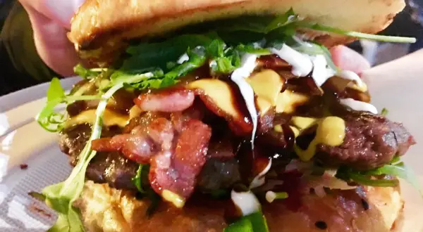 Soczysty burger z bekonem, serem, rukolą i sosami, trzymany w dłoni.
