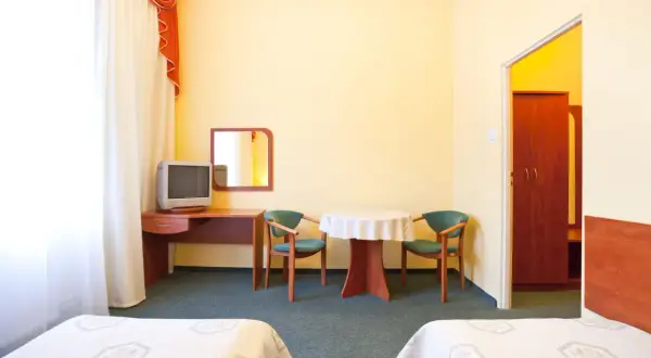 Pokój hotelowy z dwoma łóżkami, biurkiem z telewizorem, stolikiem z krzesłami i szafą.