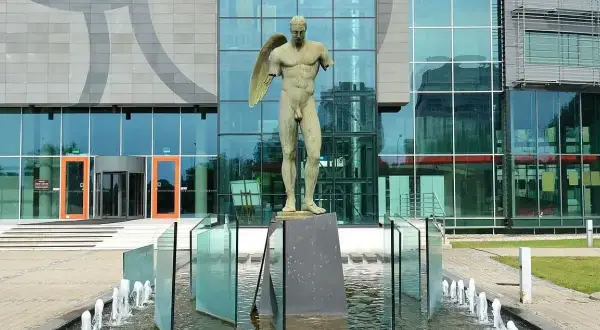 Brązowe rzeźby przedstawiające postacie ludzkie, w tym anioła, stoją przed nowoczesnym budynkiem z przeszkloną fasadą.