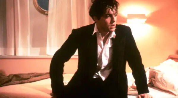 Hugh Grant w garniturze, w rozpiętej koszuli, wygląda na zamyślonego. Scena z jednego z jego filmów.