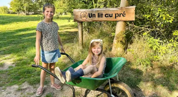 Deux petites filles profitent d'un séjour à la ferme, l'une assise dans une brouette, l'autre à côté, sous un panneau "Un lit au pré".