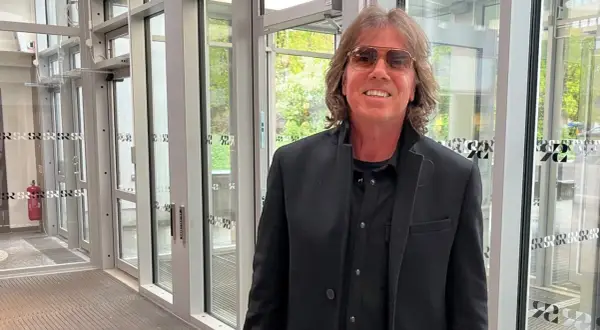 Joey Tempest, med sitt karaktäristiska hår och solglasögon, poserar framför en glasdörr.
