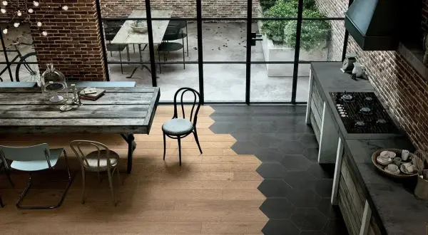 Cucina con stacco tra parquet e piastrelle esagonali nere. Tavolo rustico, sedie, mattoni a vista e grande finestra sul cortile.