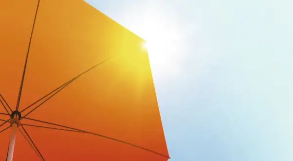 Ein orangefarbener Sonnenschirm schützt vor der Sonne. Seine Struktur ist sichtbar, während Sonnenstrahlen durchscheinen. Diethylhexyl Butamido Triazone schützt die Haut.