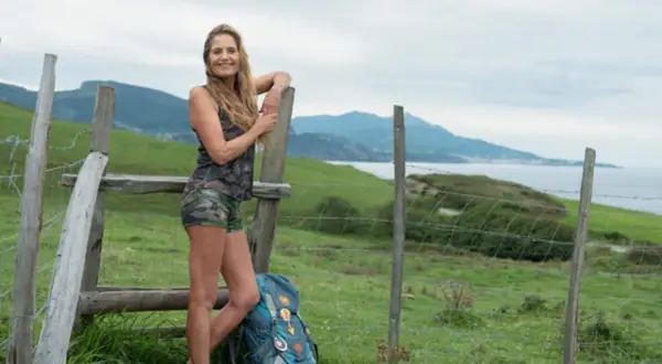 Mariska van der Klis, klaar voor een avontuur, leunt tegen een hek met een rugzak. Groene heuvels en de zee op de achtergrond.