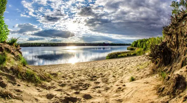 Piaskowa plaża nad jeziorem, słońce przebija się przez chmury, tworząc refleksy na wodzie. W oddali las, a po prawej stronie widoczne korzenie drzew. Idealne miejsce na wypoczynek nad pogorią zalew.