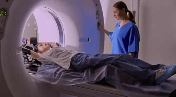 En patient ligger på en brits i en MR-scanner, medan en sjuksköterska justerar inställningarna. Mr Ryggs undersökning pågår.