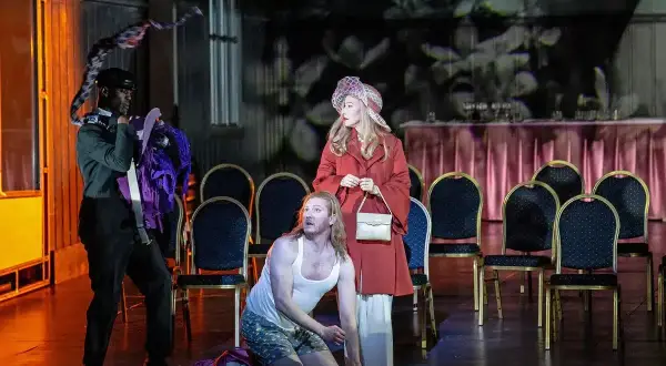 Aktorzy na scenie operetki, mężczyzna klęczy, kobieta stoi, trzeci mężczyzna trzyma kostium.
