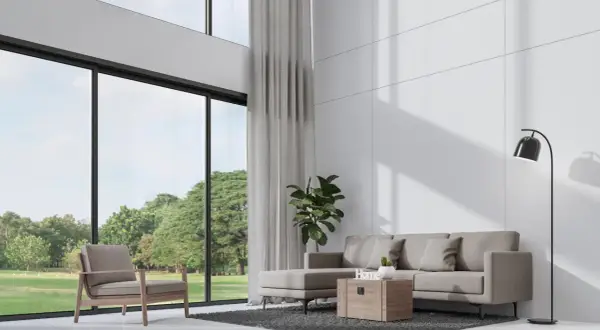 Nowoczesny salon z wysokim sufitem, szara sofa, fotel i wielkie okna z widokiem na zieleń.