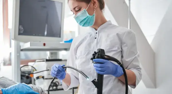 Lekarka w masce i rękawiczkach przygotowuje się do gastroskopii. Pacjent leży, a na ekranie widać obraz z kamery. Po zabiegu może pojawić się ból po gastroskopii.