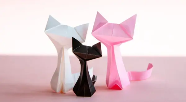 Trzy papierowe koty origami: biały, czarny i różowy, na jasnoróżowym tle.