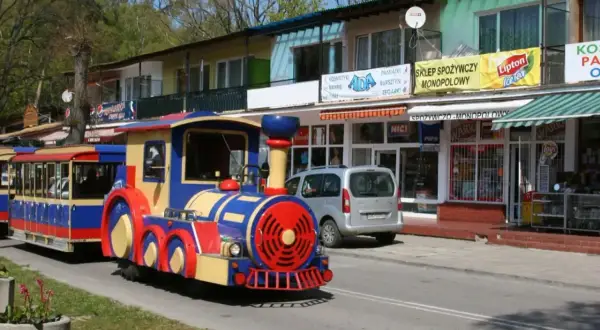 Kolorowa ciuchcia z wagonikami jedzie ulicą, zapraszając na wycieczkę, gdzie pociągiem nad morze. W tle sklepy z pamiątkami i spożywcze.