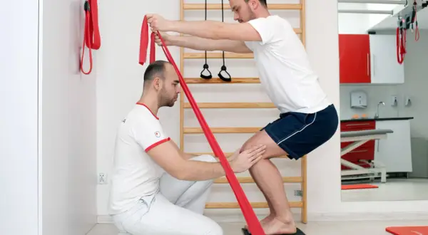 Fizjoterapeuta pomaga pacjentowi w ćwiczeniach z taśmą oporową, wspierając domową rehabilitację stawu skokowego.