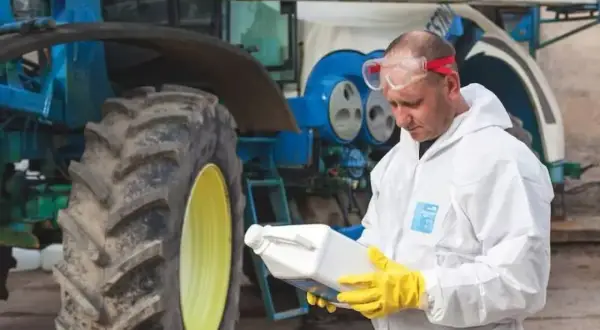 Rolnik w kombinezonie ochronnym i rękawicach trzyma butelkę z preparatem, prawdopodobnie zawierającym difenokonazol, obok traktora.