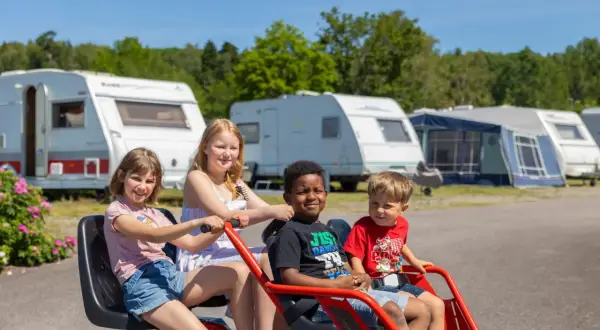 Fyra glada barn på en trampbil på Kolmården first camp. Husvagnar och träd i bakgrunden.