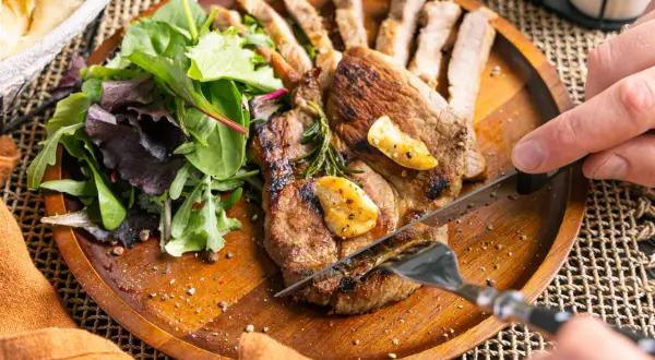 Ein saftiges Schweinesteak in der Pfanne, mit Rosmarin und Knoblauch verfeinert, wird mit einem Messer und einer Gabel angeschnitten. Dazu gibt es einen frischen Salat.