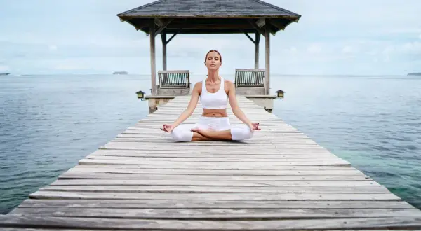 Frau in weißer Kleidung meditiert auf einem Steg am Meer. Eine Anleitung zur transzendentalen Meditation, um inneren Frieden zu finden.