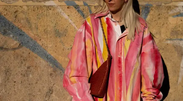 Kobieta w okularach przeciwsłonecznych i koszuli w stylu tie-dye w odcieniach różu i żółci, z brązową torebką na łańcuszku.