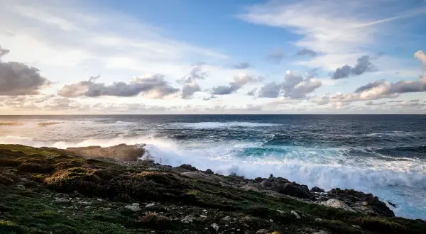 O mar bravo bate nas rochas rochosas, espumando e rugindo. O céu azul com nuvens brancas paira sobre o vasto oceano, um cenário perfeito para o portiño.