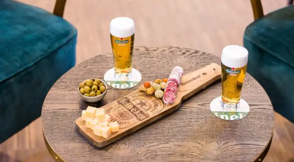 Due bicchieri di birra ale Heineken con salumi, formaggio e olive su un tavolino di legno.