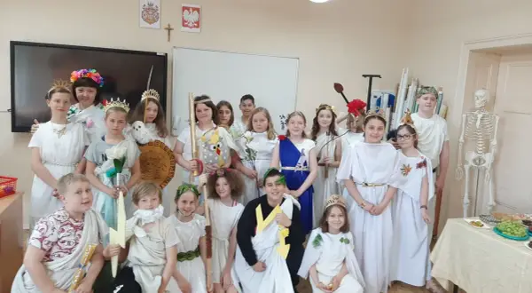 Dzieci w strojach antycznych bogów i bogiń, jak Artemida, pozują do zdjęcia. W tle szkielet i tablica.