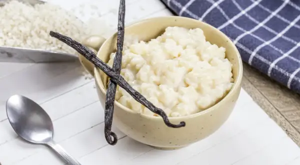 Un bol de riz au lait crémeux, garni de gousses de vanille, avec du riz brut dans une pelle à côté.