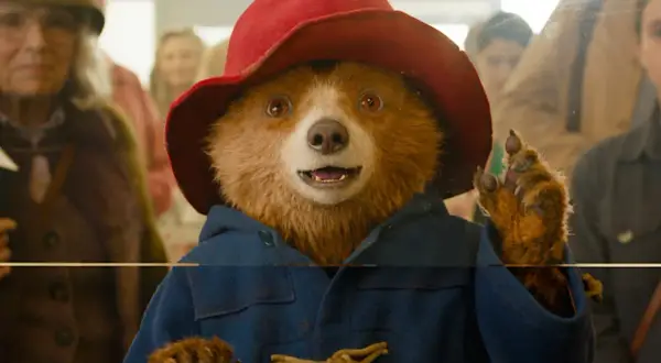 Pluszowy miś Paddington w czerwonym kapeluszu i niebieskim płaszczu, machający łapką.