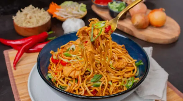 Bami Goreng mit Hühnchen, garniert mit Frühlingszwiebeln und Chili. Daneben sind Zutaten wie roter Chili, Knoblauch und Zwiebeln zu sehen.