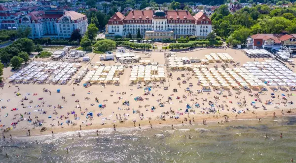Widok z lotu ptaka na zatłoczoną plażę z białymi parasolami i hotelami w tle.