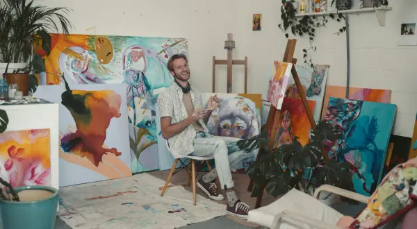 A smiling artist sits in his vibrant artist studio, surrounded by colorful canvases and plants.