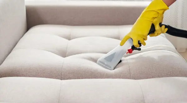 Gelbe Handschuhe führen ein Reinigungsgerät über ein helles Sofa. Tiefenreinigung für ein makelloses Sofa.