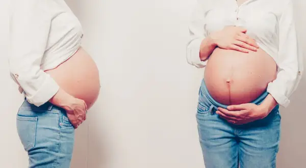 Dwa zaokrąglone brzuchy w ciąży, obie kobiety w białych koszulach i dżinsach, delikatnie dotykają swoich brzuchów.