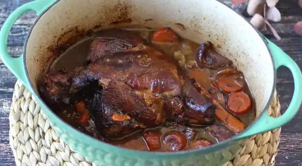 Un gigot de 7 heures mijoté dans une cocotte verte, avec des carottes et des oignons. La viande est tendre et dorée, prête à être dégustée.