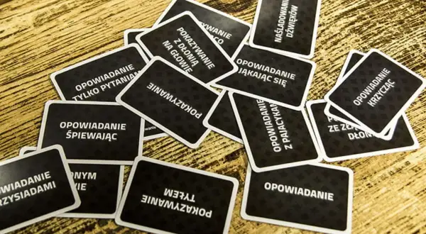 Karty do gry "Czółko" online leżą na drewnianym stole. Zadania na kartach to m.in. "Opowiadanie śpiewając", "Pokazywanie z dłonią na głowie", "Naśladowanie dźwięków".