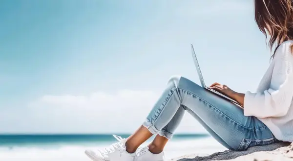 Junge Frau mit Laptop am Strand, genießt die Sonne und das Meer. Ein perfekter Ort für ihr IU Erasmus-Projekt.