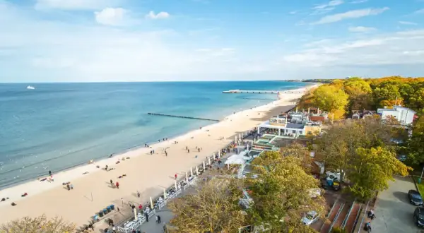 Słoneczny dzień na kołobrzeskim deptaku. Ludzie spacerują po plaży i promenadzie, ciesząc się widokiem morza i jesiennych drzew.
