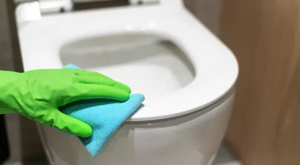Rękawica w zielonej gumie czyści deskę sedesową niebieską ściereczką. Dowiedz się, czym wyczyścić deskę sedesową, aby była higienicznie czysta.