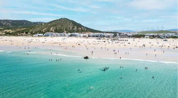 Playa de Zahara de los Atunes con gente bañándose y disfrutando del sol, con el pueblo blanco al fondo y molinos de viento en la colina.