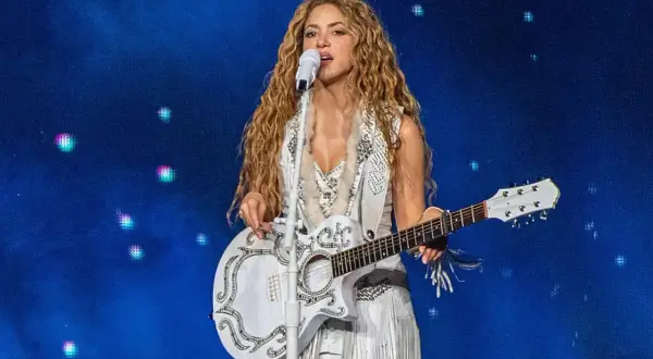 Shakira śpiewa i gra na gitarze podczas koncertu w Polsce. Wokalistka ma długie, falowane włosy i biały, zdobiony strój.