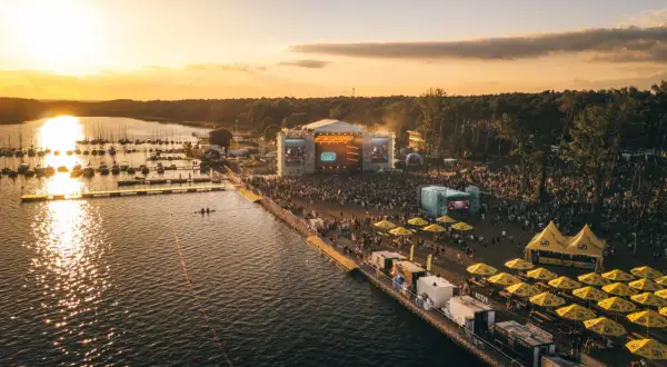Zachód słońca nad jeziorem, gdzie odbywa się festiwal. Tłumy ludzi bawią się przy scenie, a nad wodą cumują łodzie. To miejsce, gdzie rodzi się sława rap stacja.