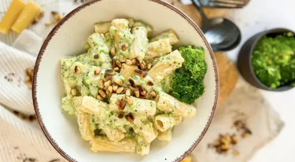 Eine Schüssel Rigatoni mit cremiger **Brokkoli Sahne Sauce** und Pinienkernen garniert. Daneben ein kleiner Napf mit gehacktem Brokkoli und Käse.