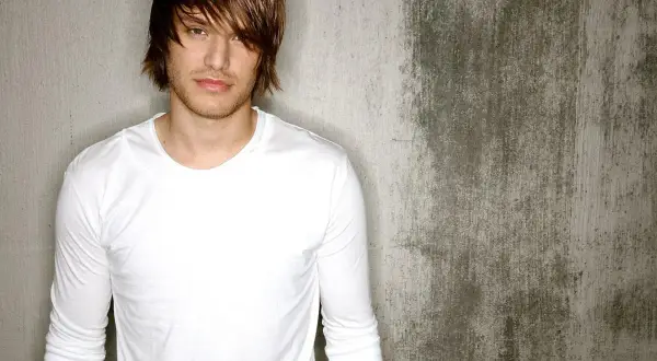 Michael Tyler, a young man with tousled brown hair and a white long-sleeved shirt, poses against a textured concrete wall.