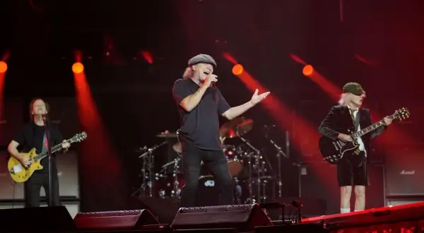 Zespół AC/DC na scenie, wokalista śpiewa, gitarzyści grają. Czerwone światła.
