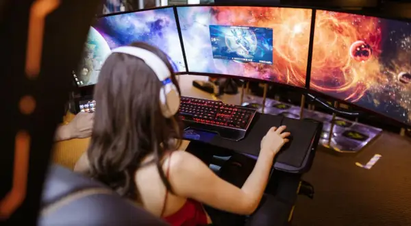 Frau mit Kopfhörern spielt an drei Monitoren. Ein "pile of shame" an Aufgaben wartet, doch jetzt zählt nur das Spiel.