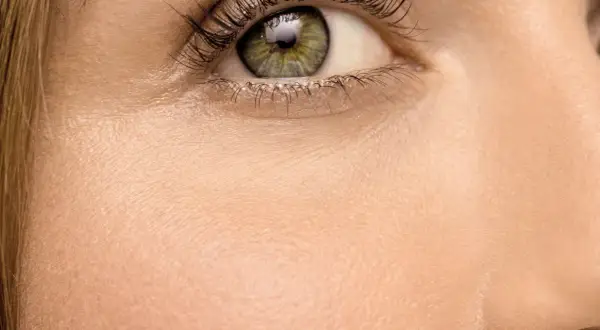 Nahaufnahme eines Auges mit grüner Iris und langen Wimpern. Die Haut wirkt blass, fast wie eine fahle Gesichtsfarbe, und zeigt feine Poren.