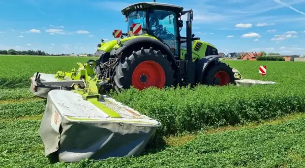 Zielony traktor z kosiarkami pracuje na polu. Rolnik zbiera plon, zastanawiając się, ile lucerny na hektar uda się uzyskać.