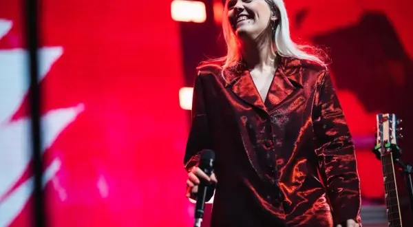 Uśmiechnięta dziewczyna pop w błyszczącej, czerwonej marynarce trzyma mikrofon na tle czerwonej sceny.