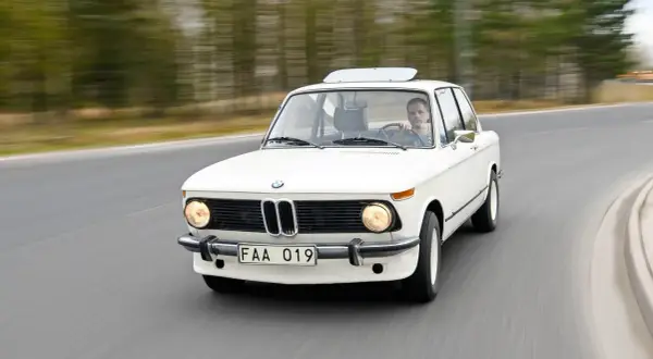 Vit BMW 70-talsklassiker kör på en väg, med en man bakom ratten.